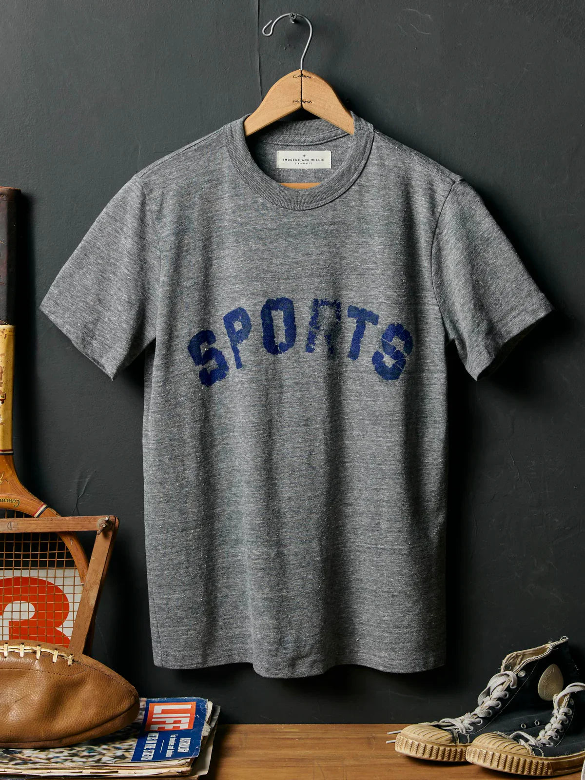Full front on hanger against dark slate wall, with distressed 'SPORTS' print, styled with football, tennis racket, Life magazine, and sneakers