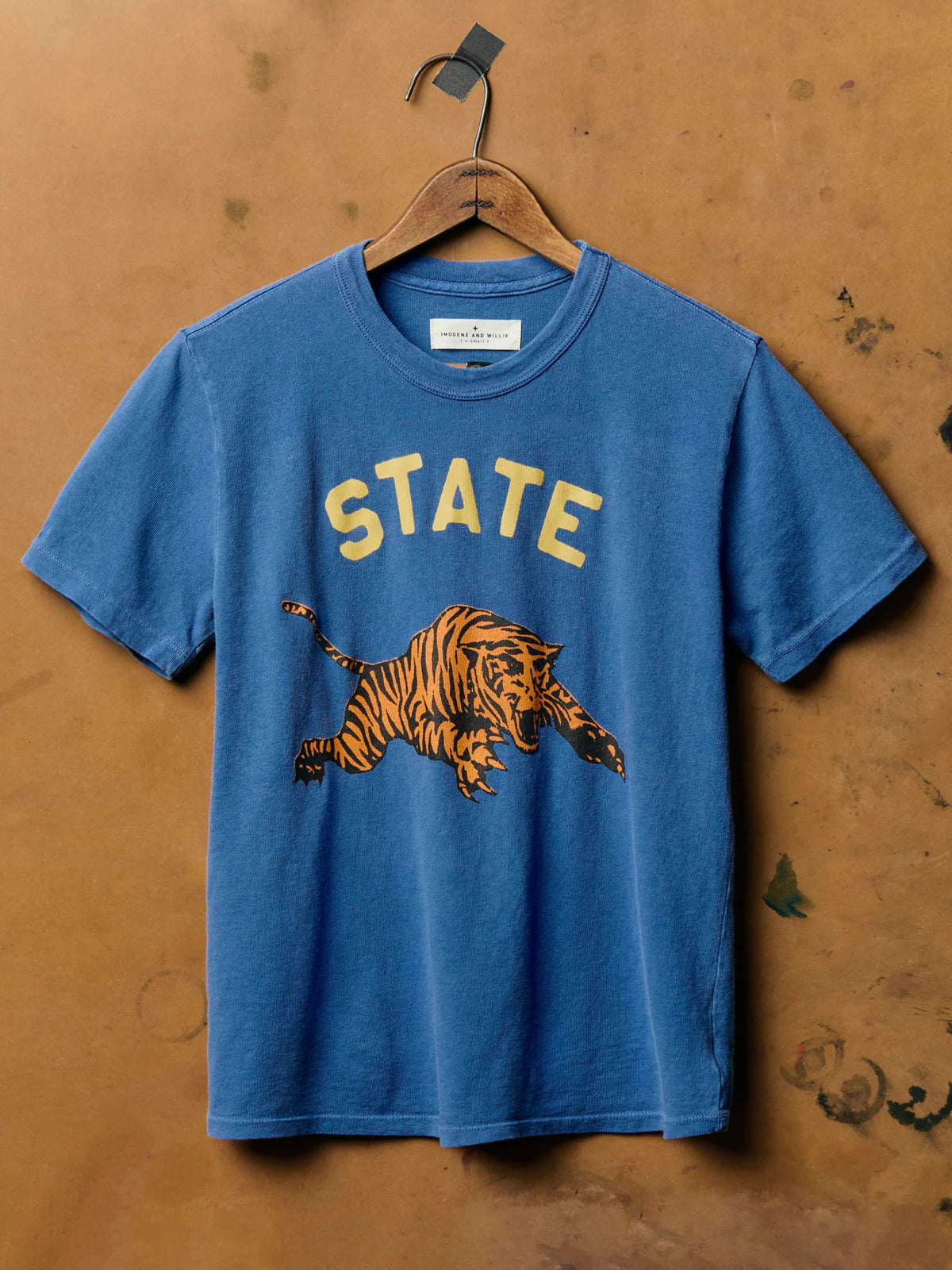 The State Tee in Blue by Imogene + Willie hanging on a wooden hanger, showing the full front with gold 'STATE' screen print text and a vintage-style leaping tiger graphic on blue cotton