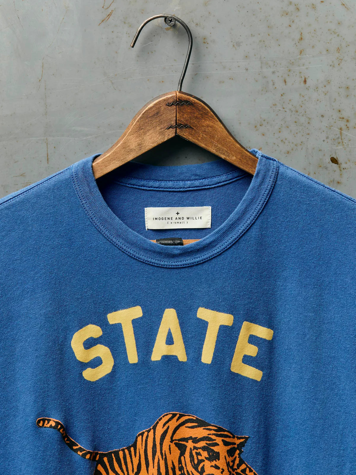 Close-up of the neckline of The State Tee in Blue showing the Imogene and Willie label, ribbed crew neck, and the top of the gold 'STATE' screen print with tiger graphic on blue cotton