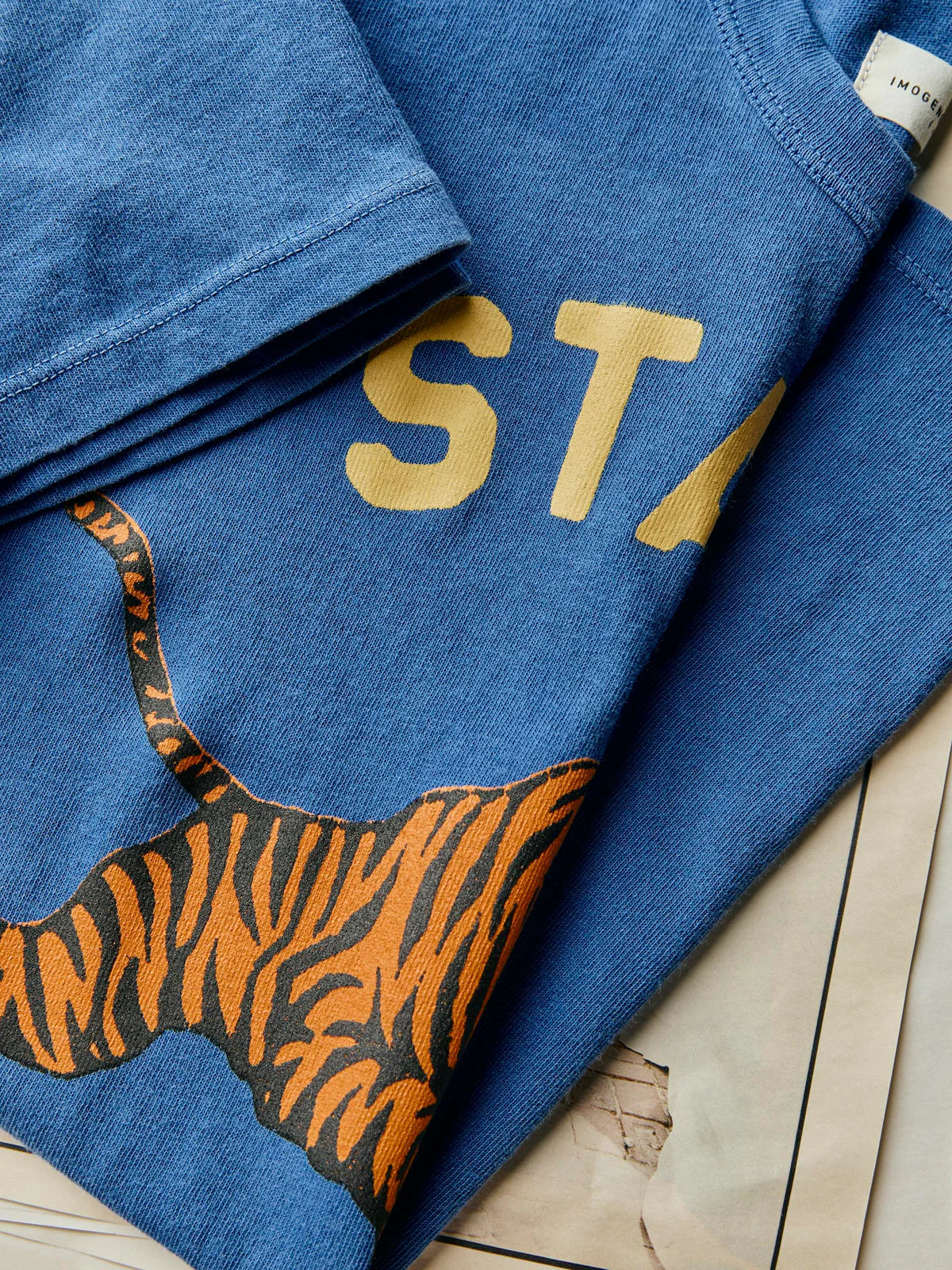 Flat lay close-up of The State Tee in Blue showing the screen-printed tiger graphic and partial 'STATE' text, highlighting the hand-drawn artwork detail and cotton fabric texture
