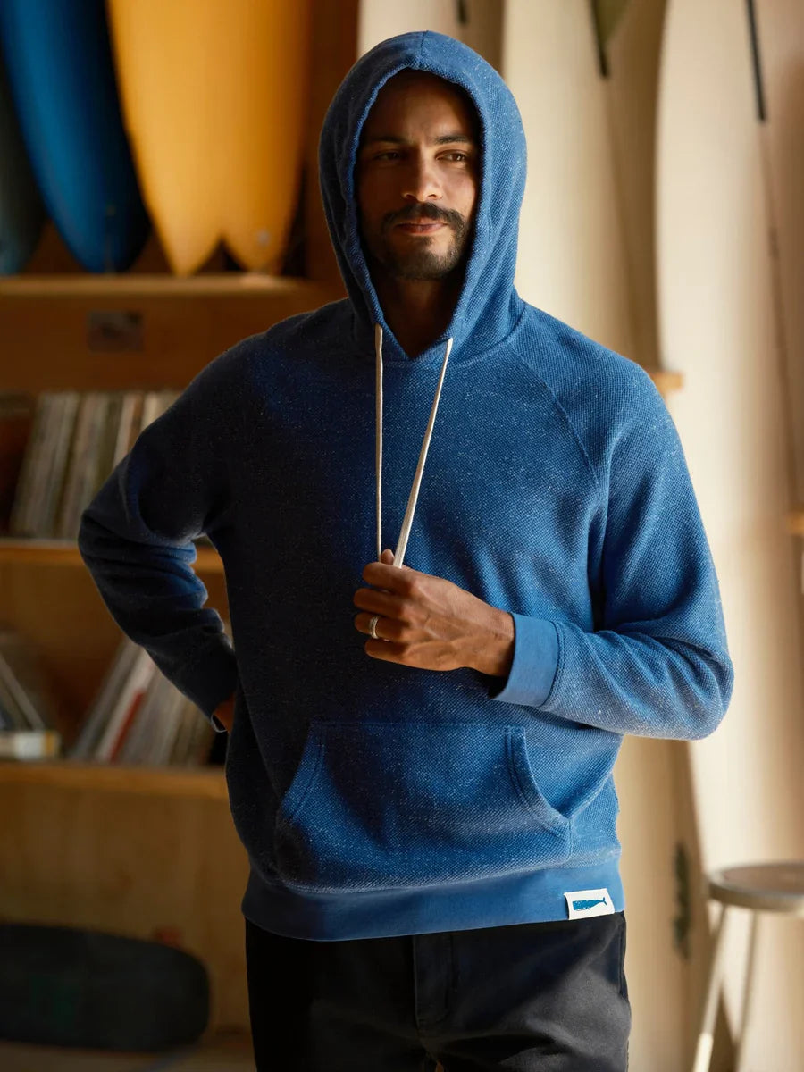 Male model wearing the Mollusk Surf Shop Whale Patch Pullover in Marine with hood up, three-quarter view showing marine blue twisted French terry texture, white drawstring, and Whale Patch label, photographed in a surf shop interior