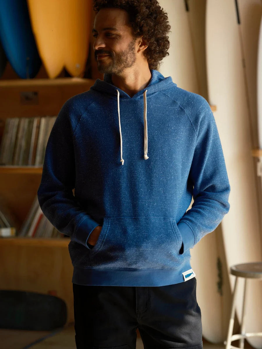 Male model wearing the Mollusk Surf Shop Whale Patch Pullover in Marine, front view showing marine blue twisted French terry hoodie with white drawstring, kangaroo pocket, and Whale Patch label on hem, photographed in a surf shop interior