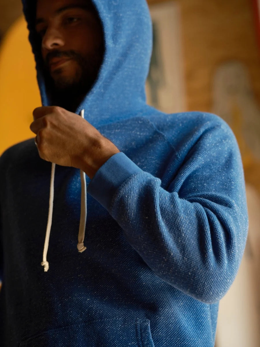 Male model wearing the Mollusk Surf Shop Whale Patch Pullover in Marine with hood up, close-up showing marine blue twisted French terry fabric, ribbed cuff, and white drawstring against a warm-toned background