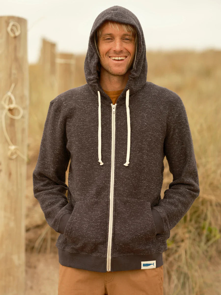 Front view of smiling male model wearing a faded navy garment-dyed zip-up hoodie with hood up and hands in kangaroo patch pockets, showing the full zip with cream pull, cream drawstring, and Olde Whale woven patch at the hem, styled with tan pants against a coastal dune background.
