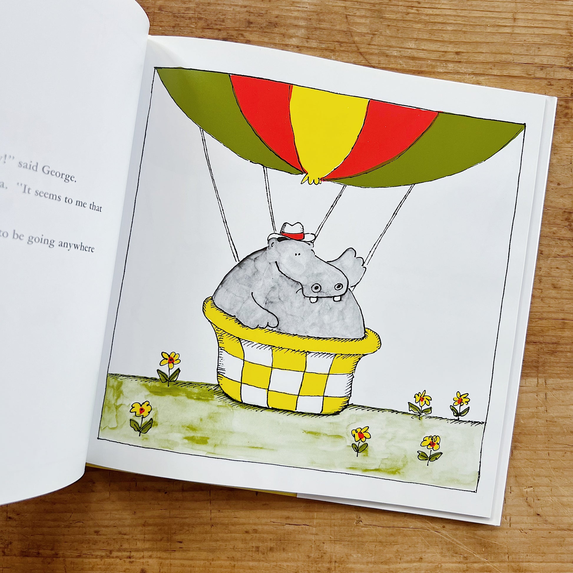 Interior spread of George and Martha from the story "The Flying Machine," showing a full-page color illustration of George the hippo wearing a white fedora, crammed into a yellow and white checkered wicker basket beneath a striped green, red, and yellow hot air balloon hovering just above a grassy field dotted with small yellow flowers, with partial text on the left page reading "said George. It seems to me that to be going anywhere," displayed on a warm wood surface.
