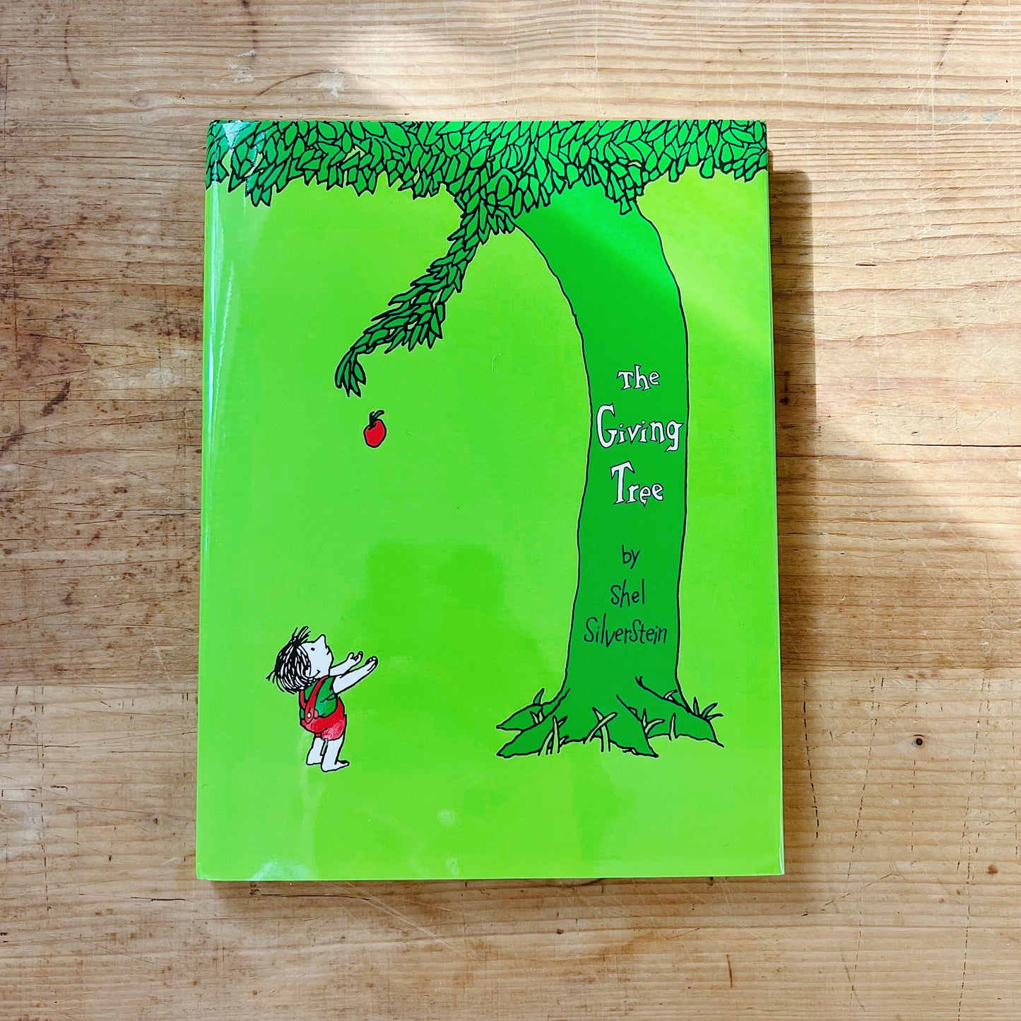 Front cover of The Giving Tree by Shel Silverstein, featuring a bright lime green background with a black-and-green ink illustration of a large leafy tree arching to the left, one branch extended with a single red apple dropping, and a small boy in a red outfit standing below with arms outstretched to catch it, the title in white hand-lettered text on the tree trunk and the author's name below, displayed flat on a warm wood surface.
