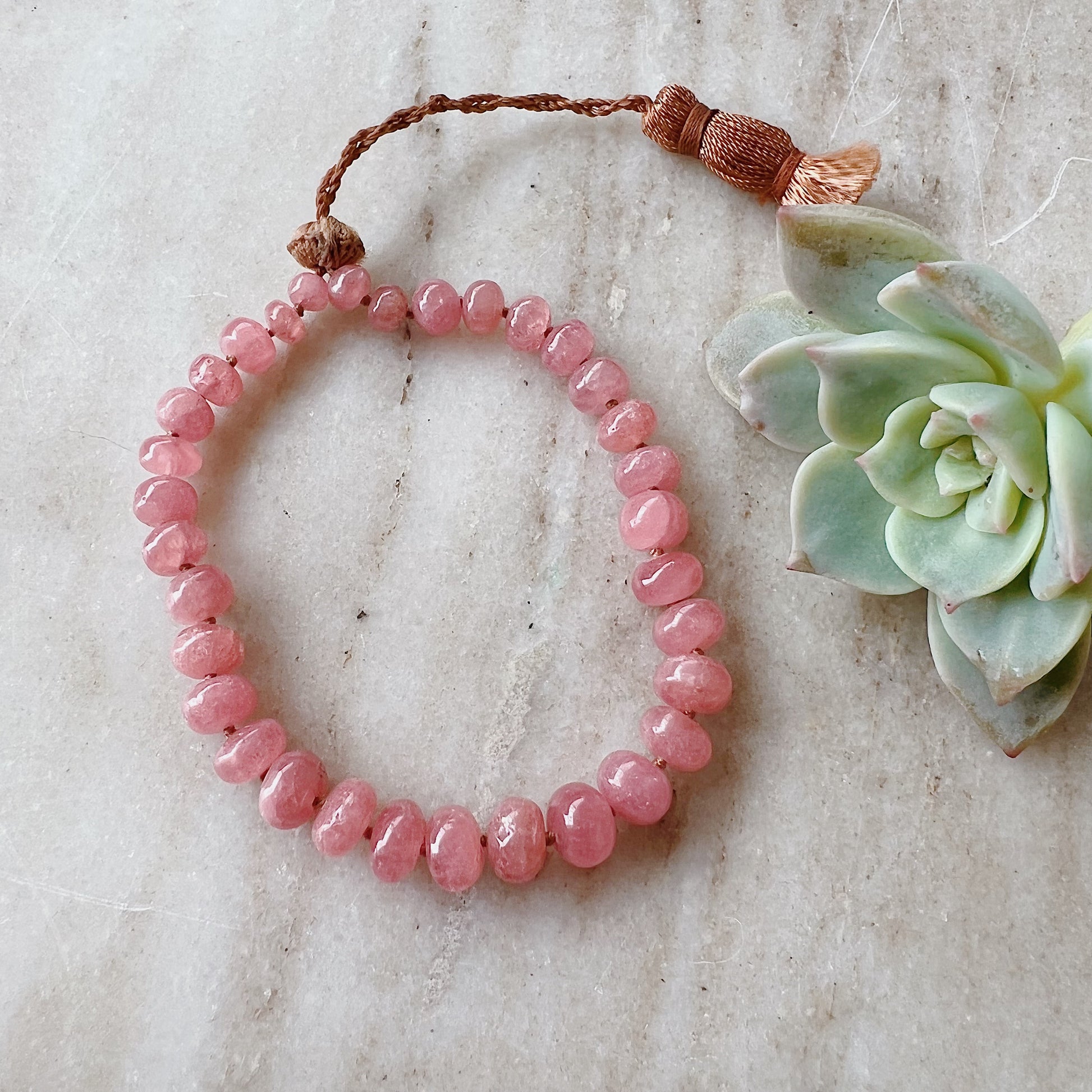 A hand-knotted waxed silk cord bracelet with pink rhodochrosite beads and an adjustable tassel closure, displayed on a marble surface next to a succulent plant.