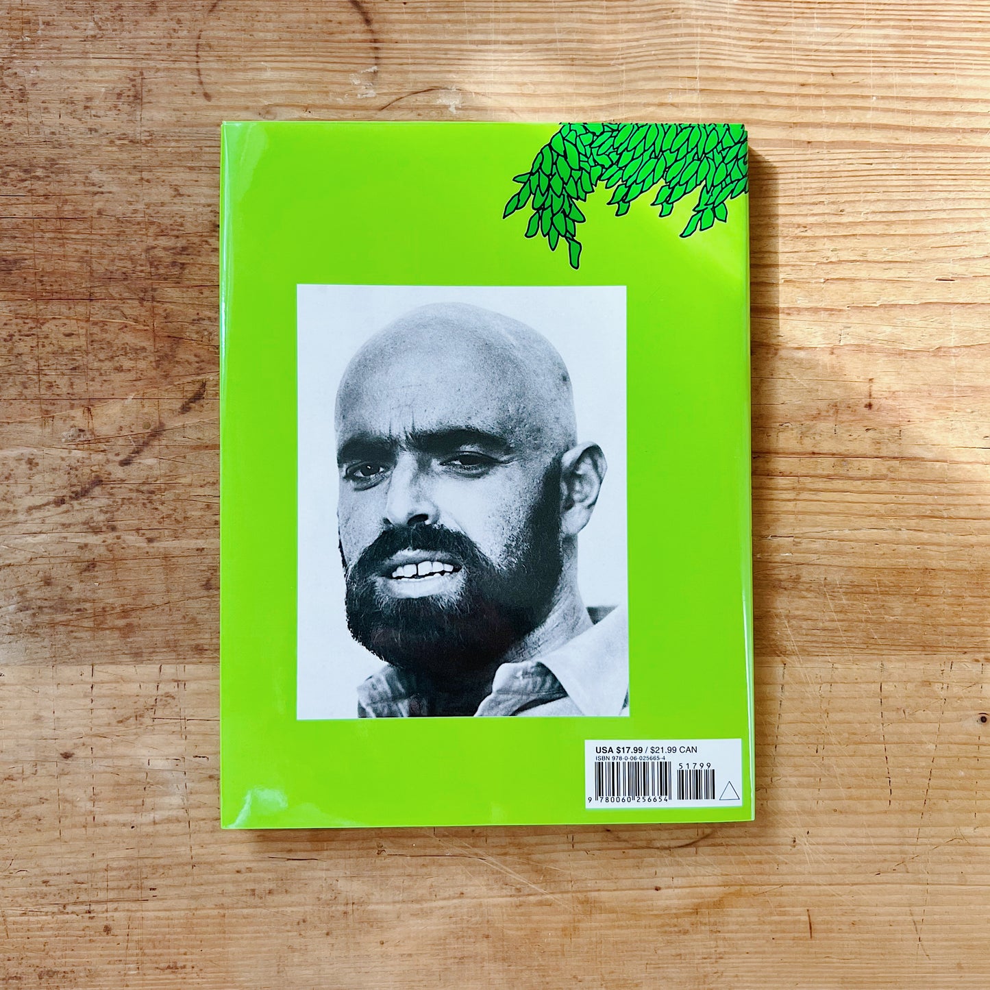Back cover of The Giving Tree on a bright lime green background with a partial green ink illustration of the tree's leafy canopy in the upper right corner, a large black-and-white author photograph of Shel Silverstein — a bald man with a dark beard and intense gaze — centered on the cover, with a barcode and retail price of $17.99 USD in the lower right corner, displayed flat on a warm wood surface.
