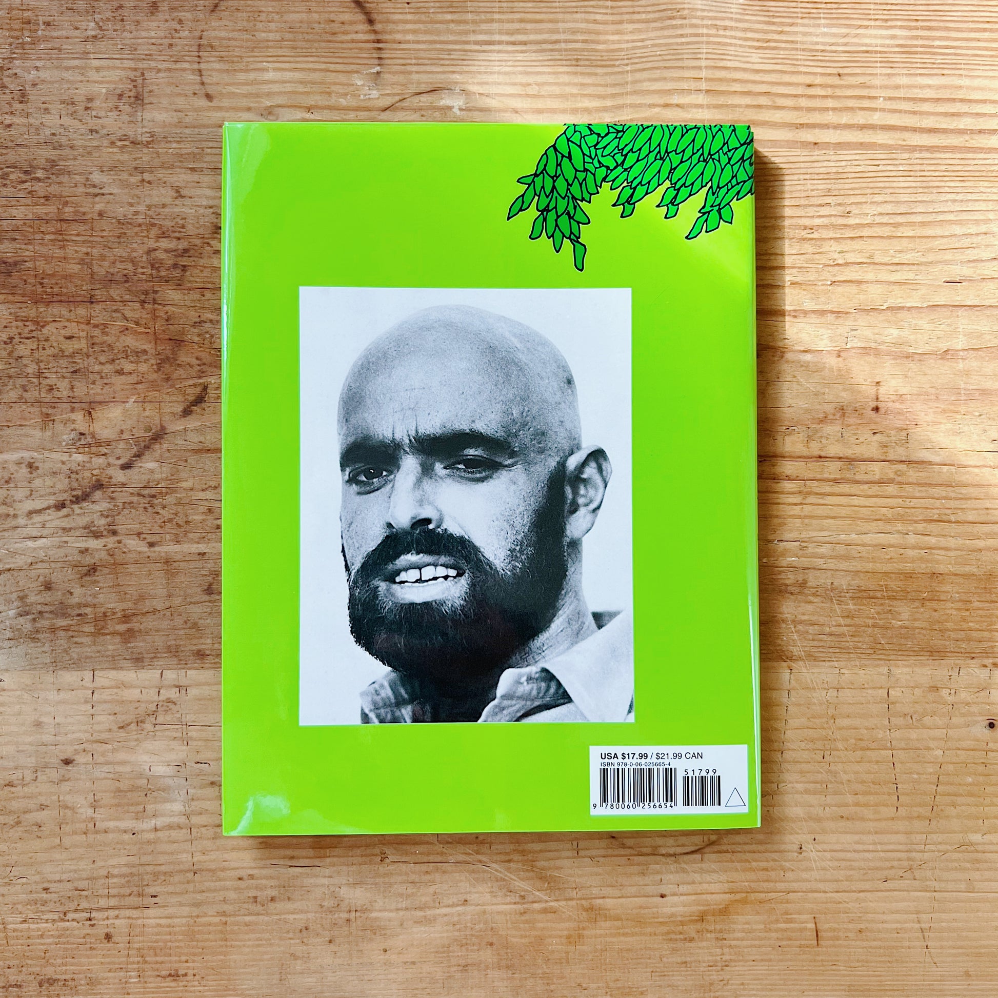 Back cover of The Giving Tree on a bright lime green background with a partial green ink illustration of the tree's leafy canopy in the upper right corner, a large black-and-white author photograph of Shel Silverstein — a bald man with a dark beard and intense gaze — centered on the cover, with a barcode and retail price of $17.99 USD in the lower right corner, displayed flat on a warm wood surface.
