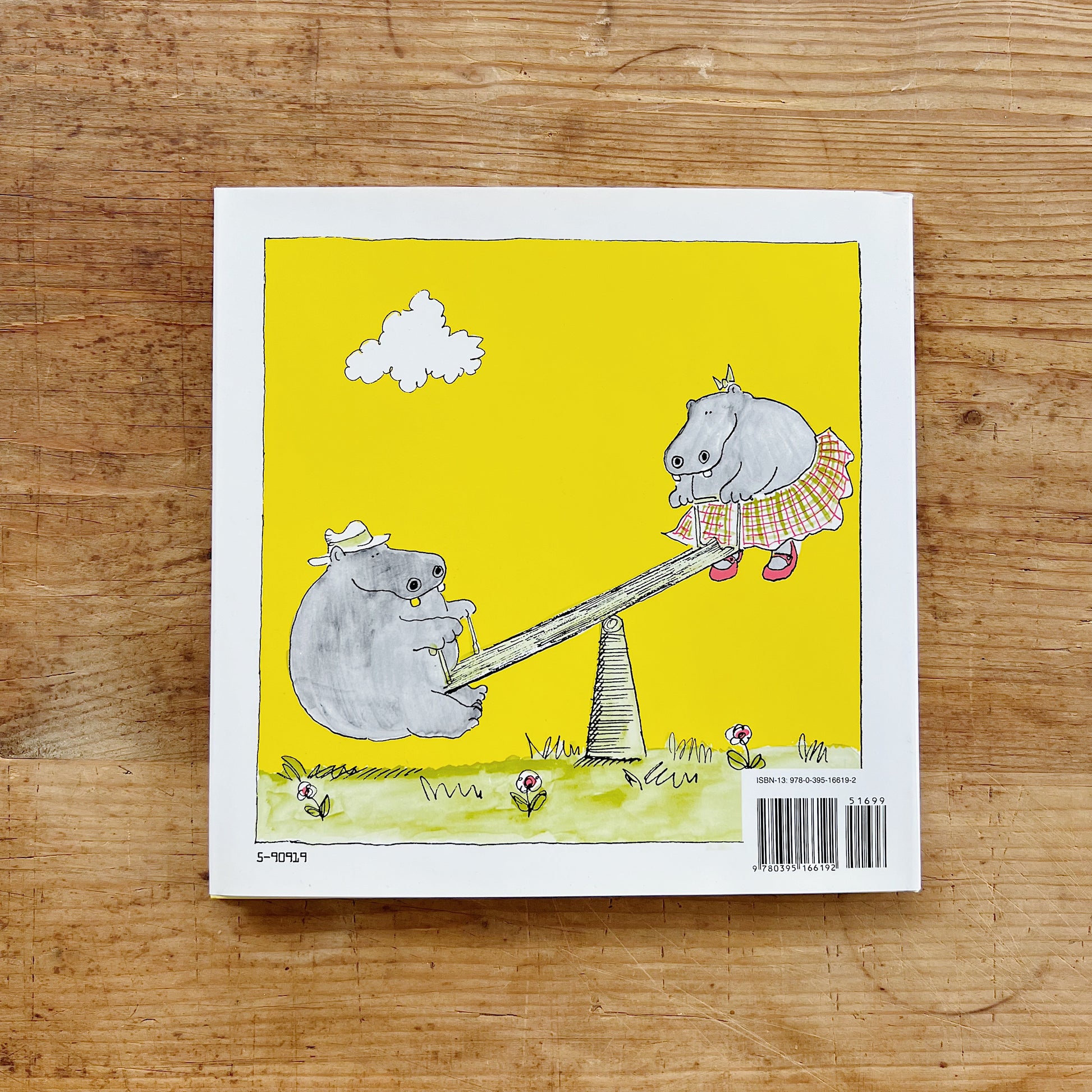 Back cover of George and Martha on a bright yellow background, showing a color illustration of George the hippo in a white hat sitting on the low end of a seesaw while Martha the hippo in a plaid skirt and red shoes sits elevated on the high end, with small flowers in the grass below and a white cloud overhead, with a barcode and retail price of $16.99 in the lower right corner, displayed flat on a warm wood surface.
