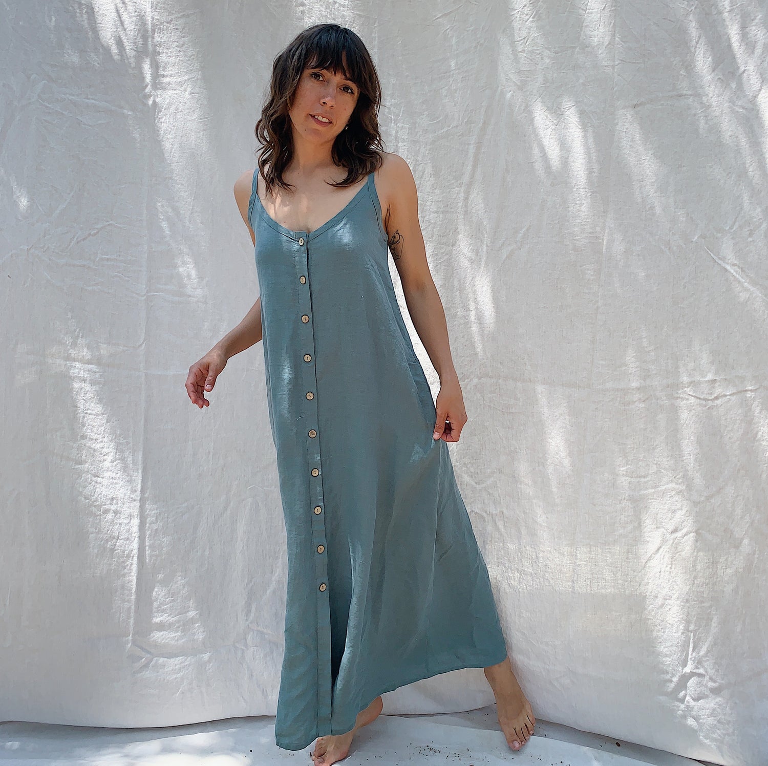 Woman with brown hair wearing a long sage green linen dress with buttons down the front on a canvas background.