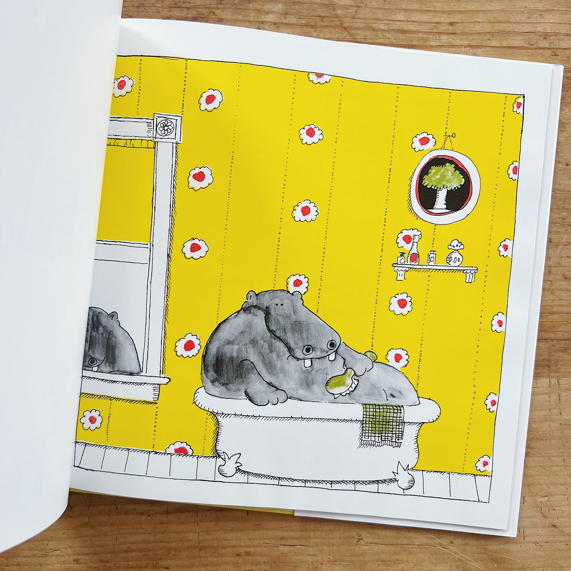 Interior spread of George and Martha from the story "The Tub," showing a full-page color illustration of Martha the hippo squeezed into a white clawfoot bathtub in a bright yellow floral-wallpapered bathroom, holding a green sponge, with a round porthole window showing a tree, bath products on a wall shelf, and two rubber ducks on the floor, displayed on a warm wood surface.
