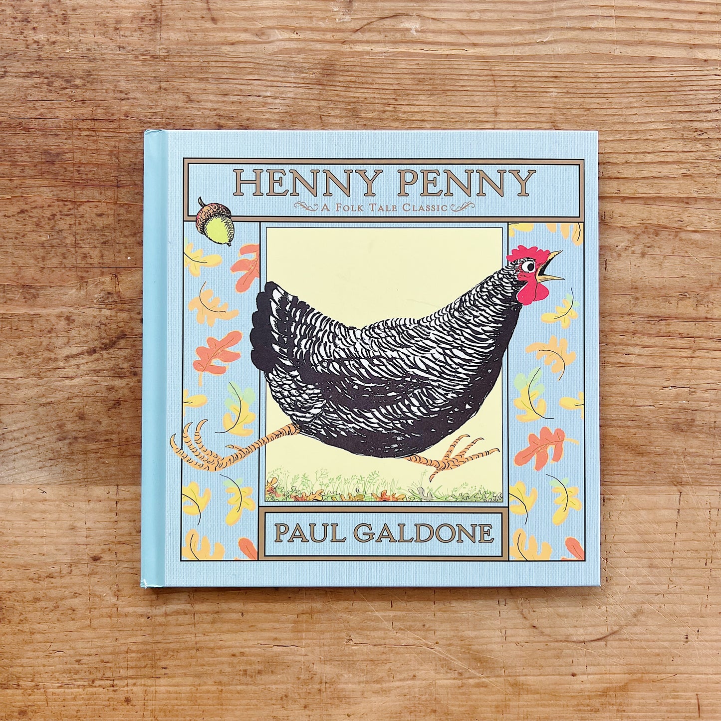 Front cover of Henny Penny by Paul Galdone, featuring a powder blue linen-textured cover with a decorative gold-ruled border and colorful autumn leaf motifs. A central framed panel shows a large barred black-and-white hen with a red comb and open beak, standing in a grassy field with a falling acorn visible in the upper left corner. The title is printed in gold serif lettering at the top and

