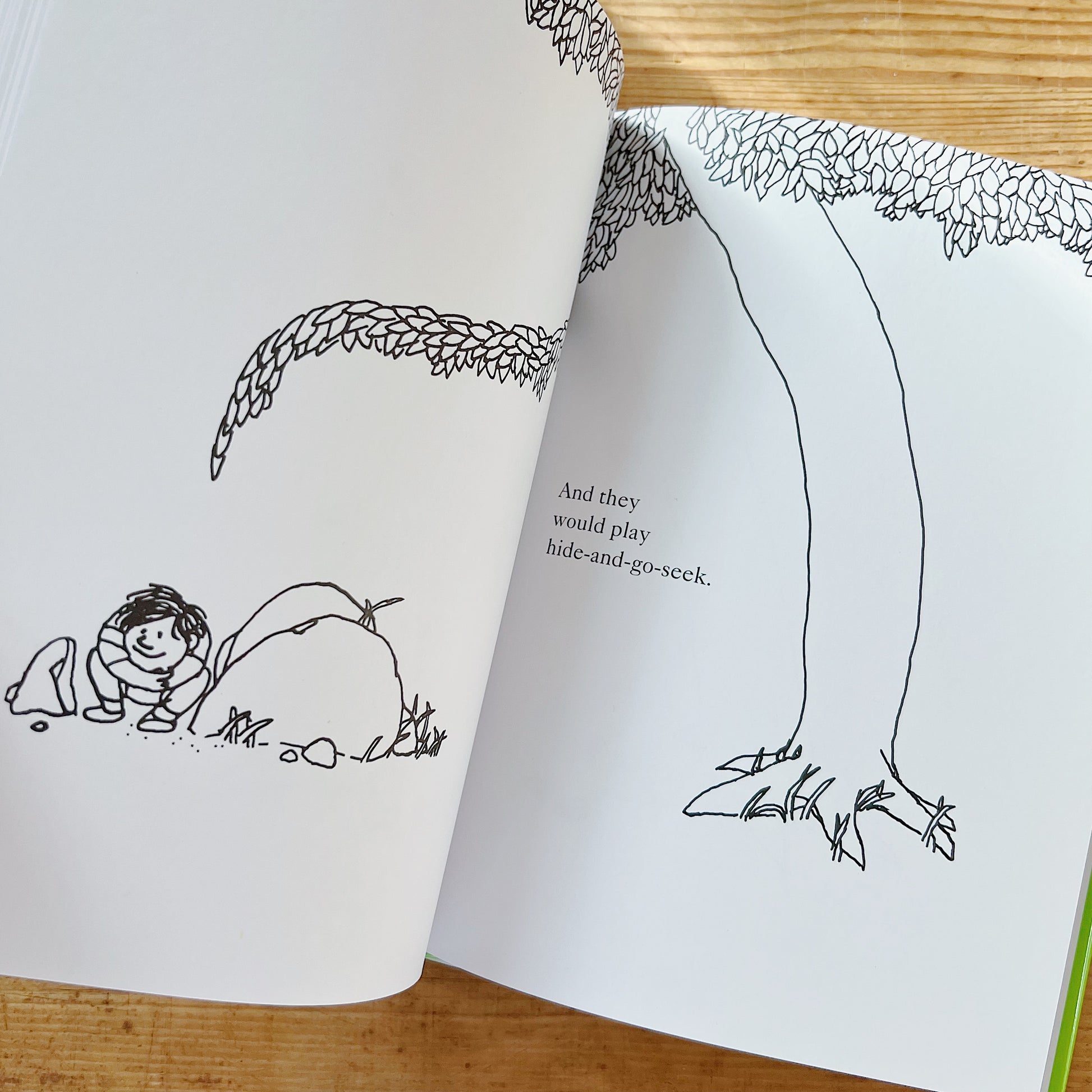Open interior spread of The Giving Tree showing two pages: on the left, a black-and-white line illustration of the young boy crouching behind a small mound of dirt at the base of the tree, smiling as he hides; on the right, the tree's tall trunk and leafy canopy with the text "And they would play hide-and-go-seek," displayed on a warm wood surface.
