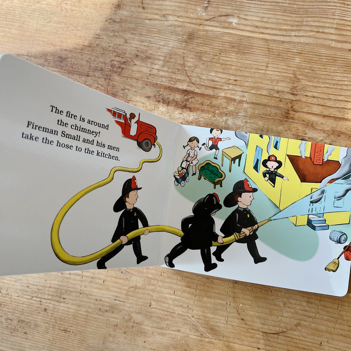 Interior spread of The Little Fire Engine showing Fireman Small and two firefighters carrying a yellow hose toward a smoking yellow house, beside text reading "The fire is around the chimney! Fireman Small and his men take the hose to the kitchen"
