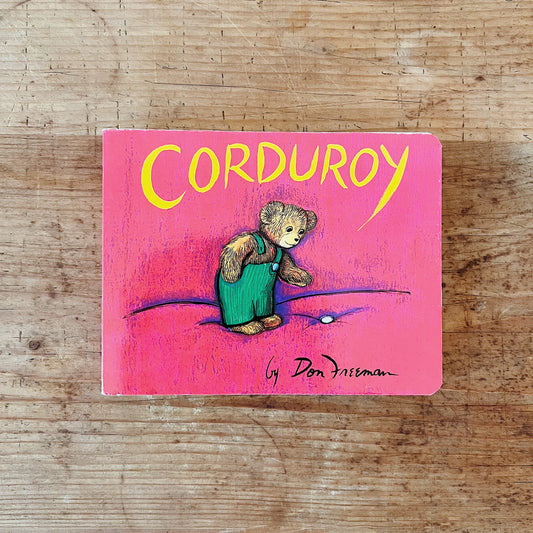 Cover of Corduroy board book by Don Freeman, featuring a red background with the title in large yellow lettering. Corduroy, a small brown teddy bear in green overalls, stands looking down at a single white button on the ground, his expression wistful.

