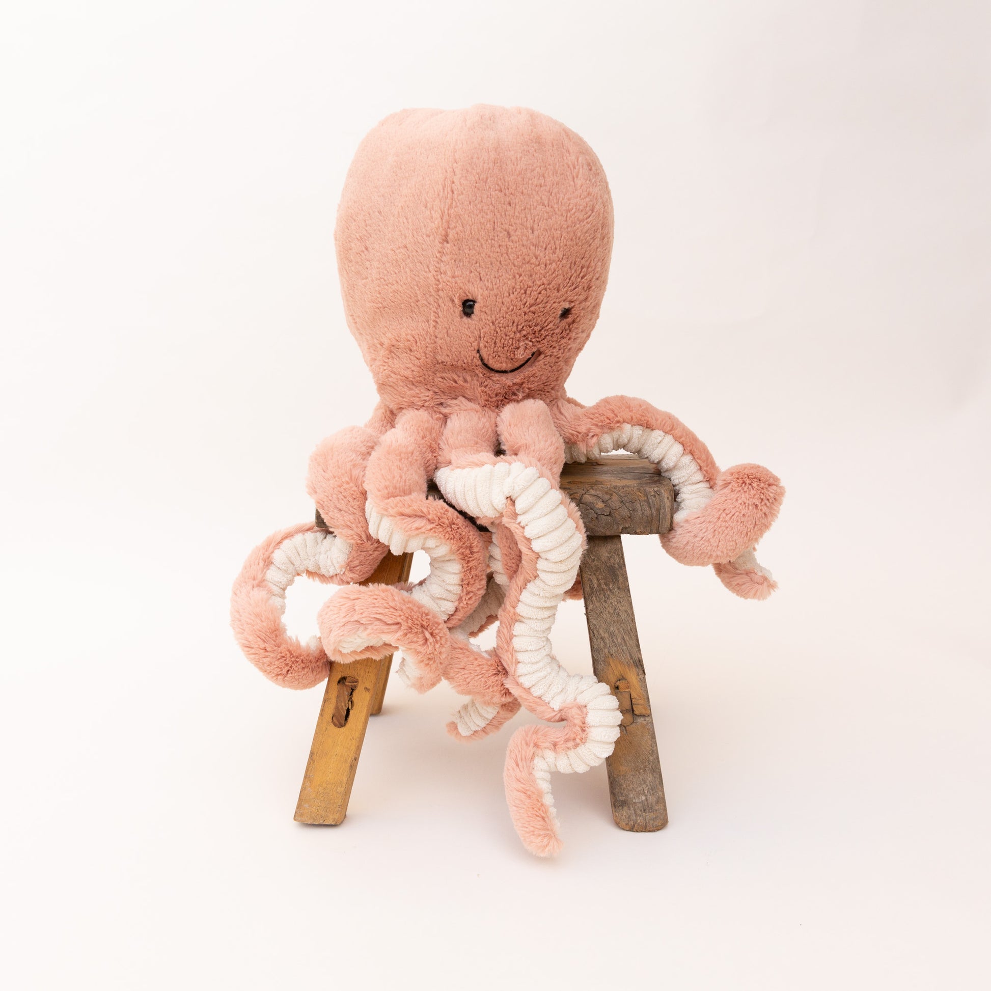 Jellycat Large Odell Octopus plush toy in blush pink, upright seated on a wooden stool showing a smiling face and eight dangling tentacles with cream sucker details on white background