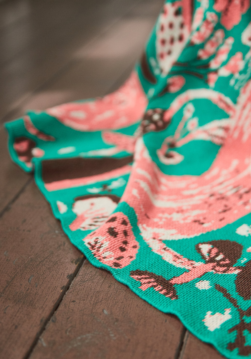 Mushroom Emerald blanket draped on a wooden floor, showing pink mushrooms and woodland motifs on green knit.
