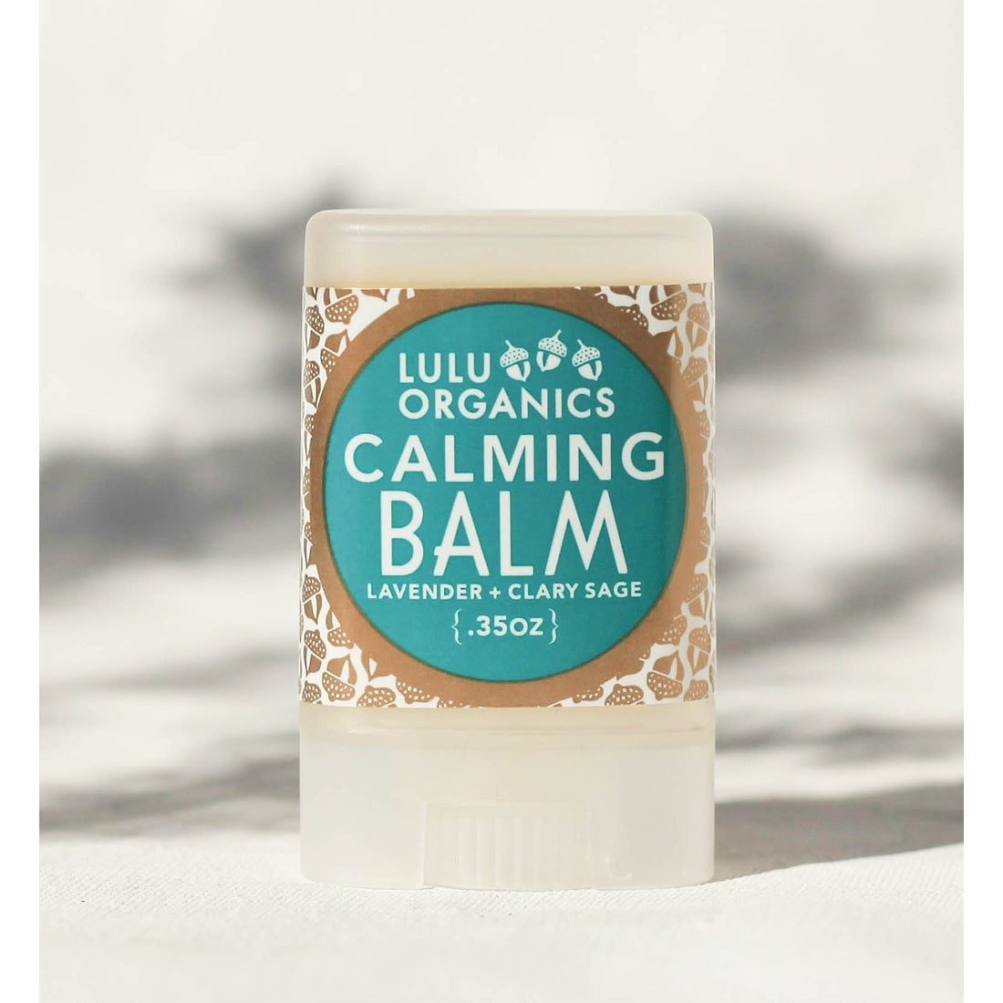 A stick of Lulu Organics Calming Balm with Lavender and Clary Sage, displayed vertically with a label showing the product name and scent, against a blurred background.