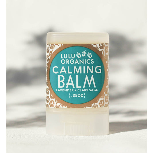 A stick of Lulu Organics Calming Balm with Lavender and Clary Sage, displayed vertically with a label showing the product name and scent, against a blurred background.