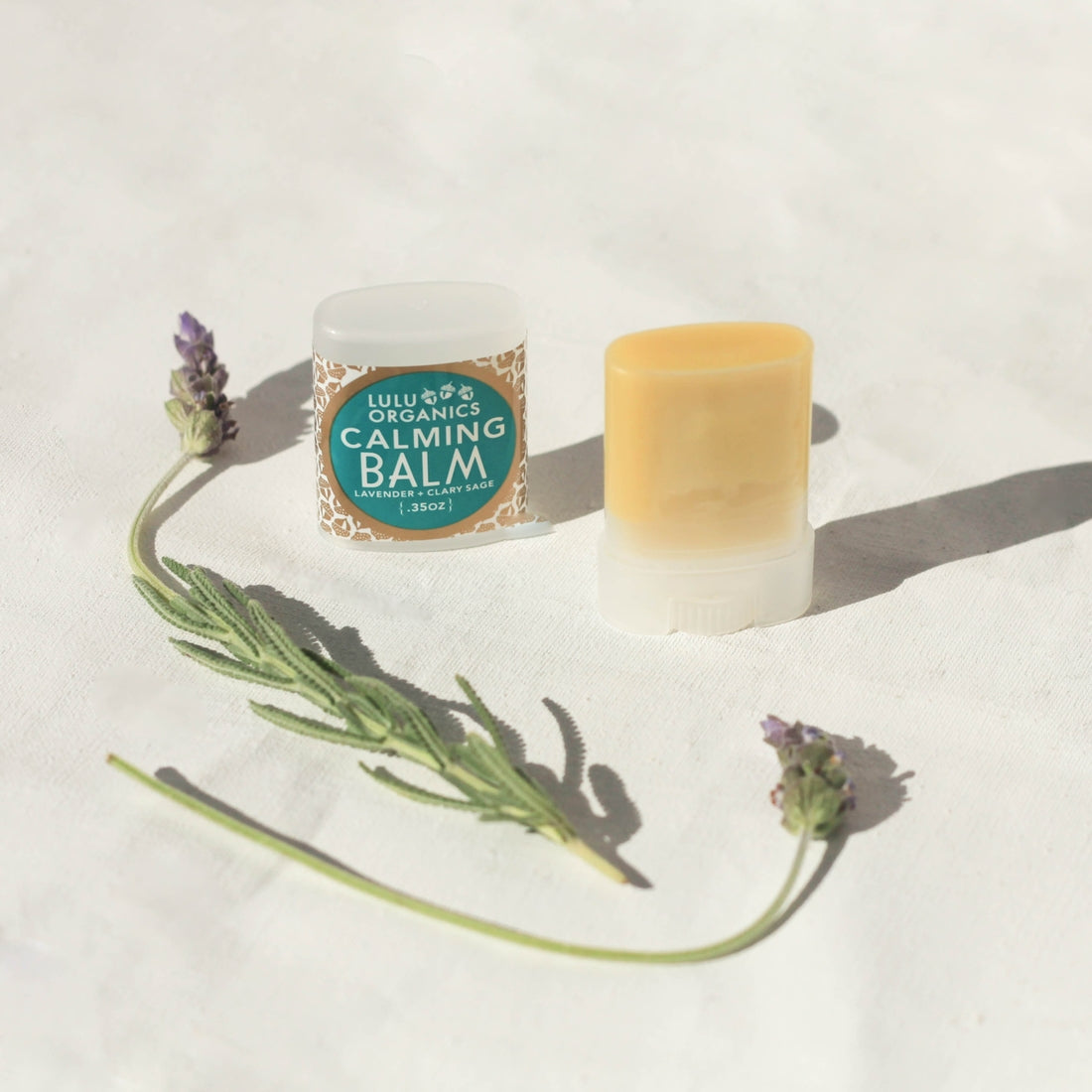 Lulu Organics Calming Balm container and open balm stick displayed with fresh lavender sprigs on a white surface in natural sunlight