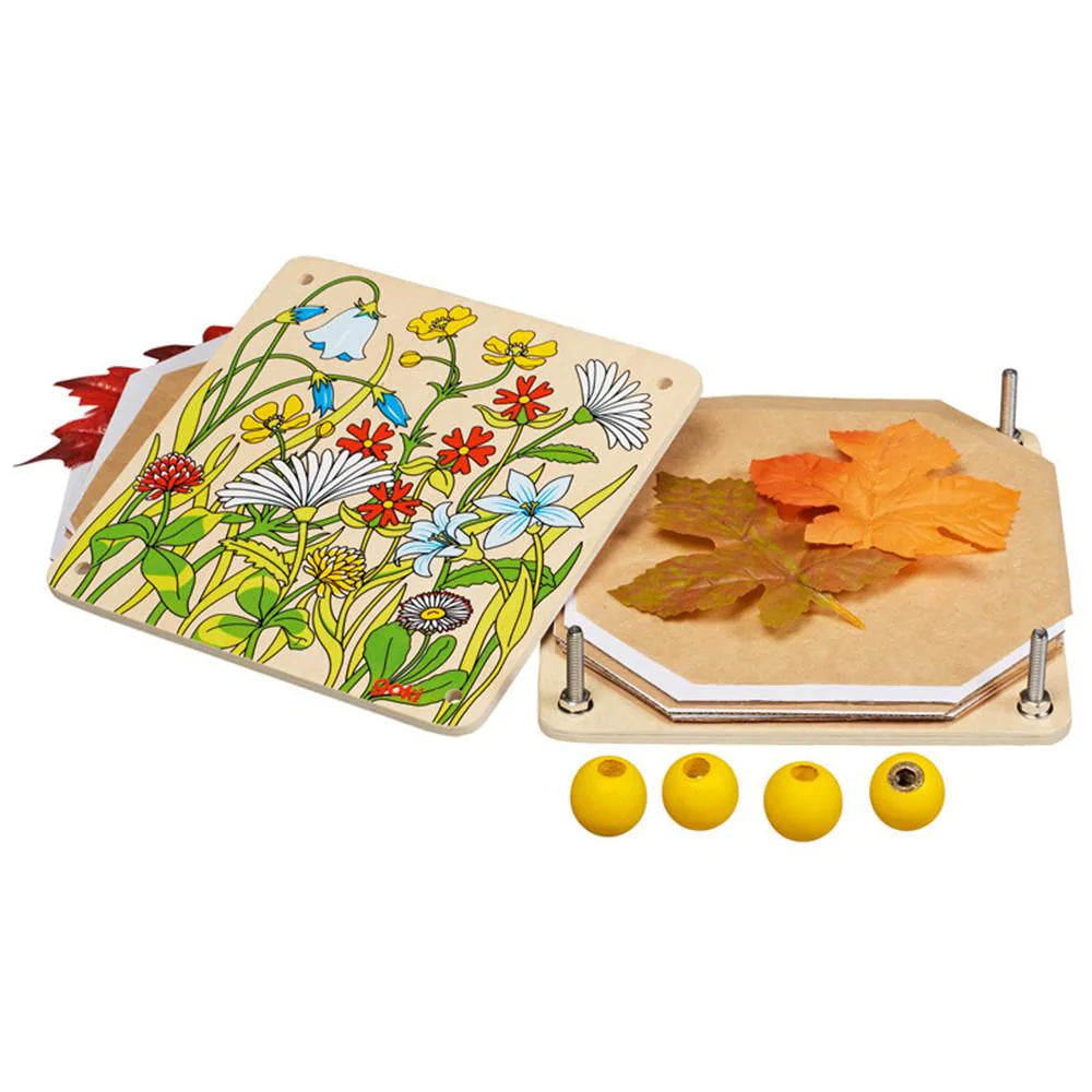  Goki Flower Press open and disassembled on a white background, showing the illustrated wooden top plate with a wildflower meadow design, the hexagonal wooden base plate loaded with autumn leaves and paper inserts, and four yellow wooden screw knobs