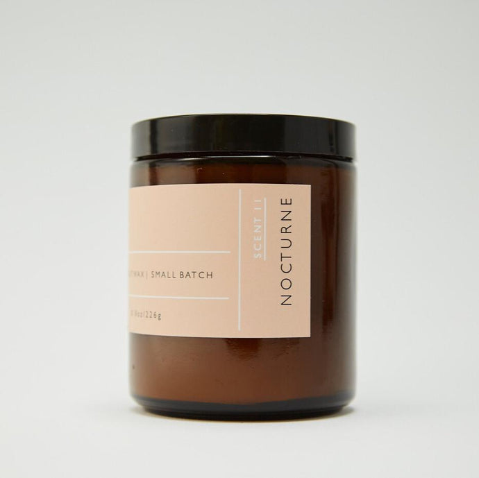 Side view of Roen's Nocturne scented candle in an amber brown glass jar with a black lid and pink label.